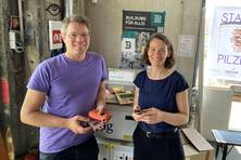 Foto von einem Mann und einer Frau in einer Werkstatt. Der Mann hält zwei Handys in der Hand. Beide lächeln in die Kamera.