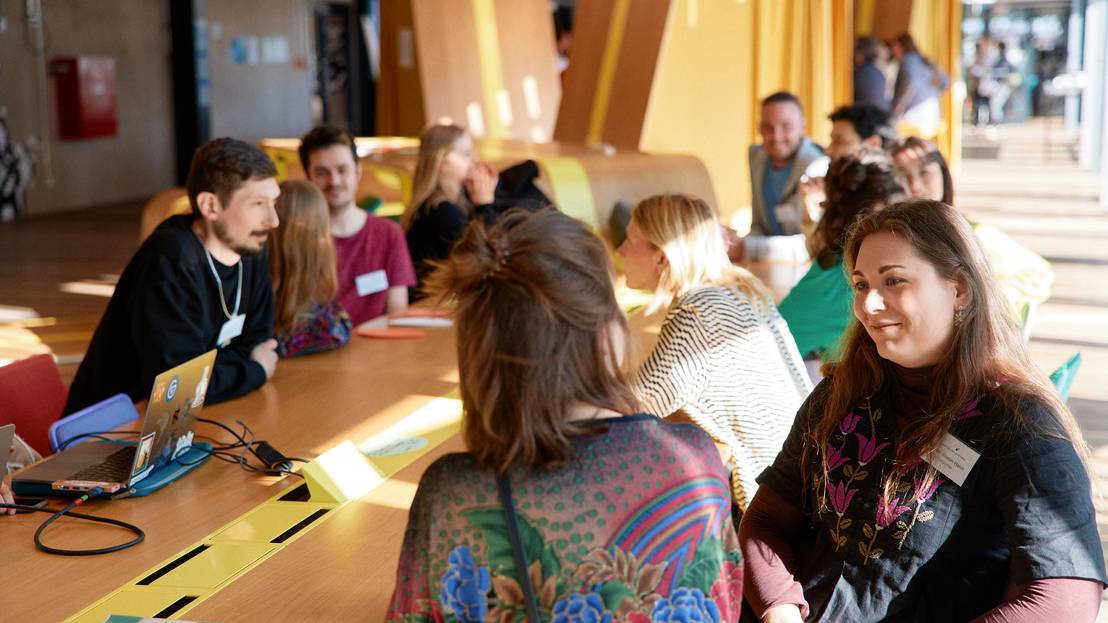 Eine Gruppe von Menschen sitzt an einem langen Tisch. Jeweils 2 Personen tauschen sich miteinander aus.